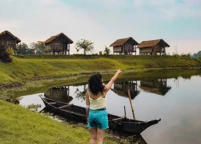 A tourist pointing towards traditional riverside eco-cottages on Majuli Island, Assam.